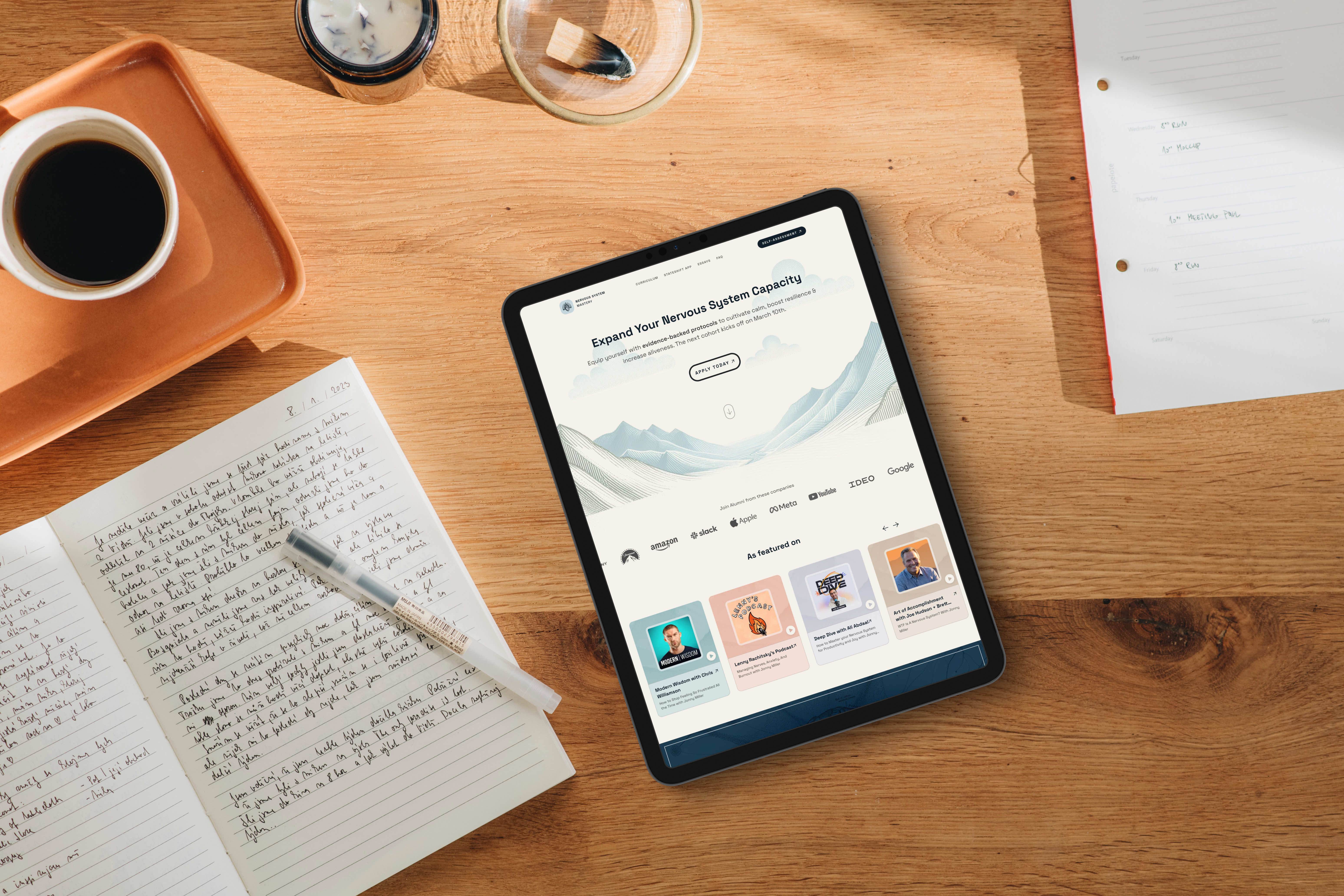 Startseite von Nervous System Mastery auf einem Tablet, das auf einem Holzschreibtisch neben einem Kaffee, einer Kerze und einem aufgeschlagenen handgeschriebenen Notizbuch liegt.
